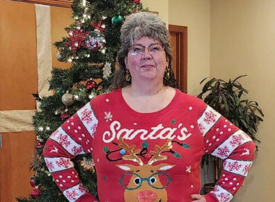 Branch Manager Dede Hoerr stands in front of a charity Christmas tree wearing a Christmas sweater.