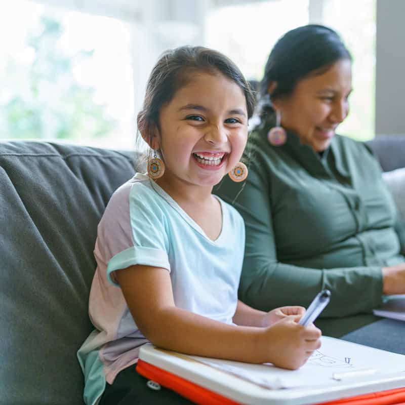 Portrait of a cute elementary age Native American girl laughing and looking directly at the camera while sitting on the couch and coloring. The little girl's mom is sitting next to her and is using a laptop computer.