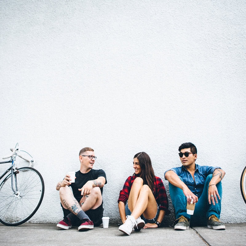 A mixed-race group of three young adults pauses for a break on their bike commute. They rest with their fixie bicycles to laugh and talk in an urban setting while enjoying coffee. They sit together on a sidewalk and lean against a concrete wall. Horizontal with copy space.